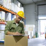 serious-male-logistic-engineer-hardhat-overall-standing-stack-boxes-completing-documents-copy-space-blue-collar-labor-concept_74855-14265